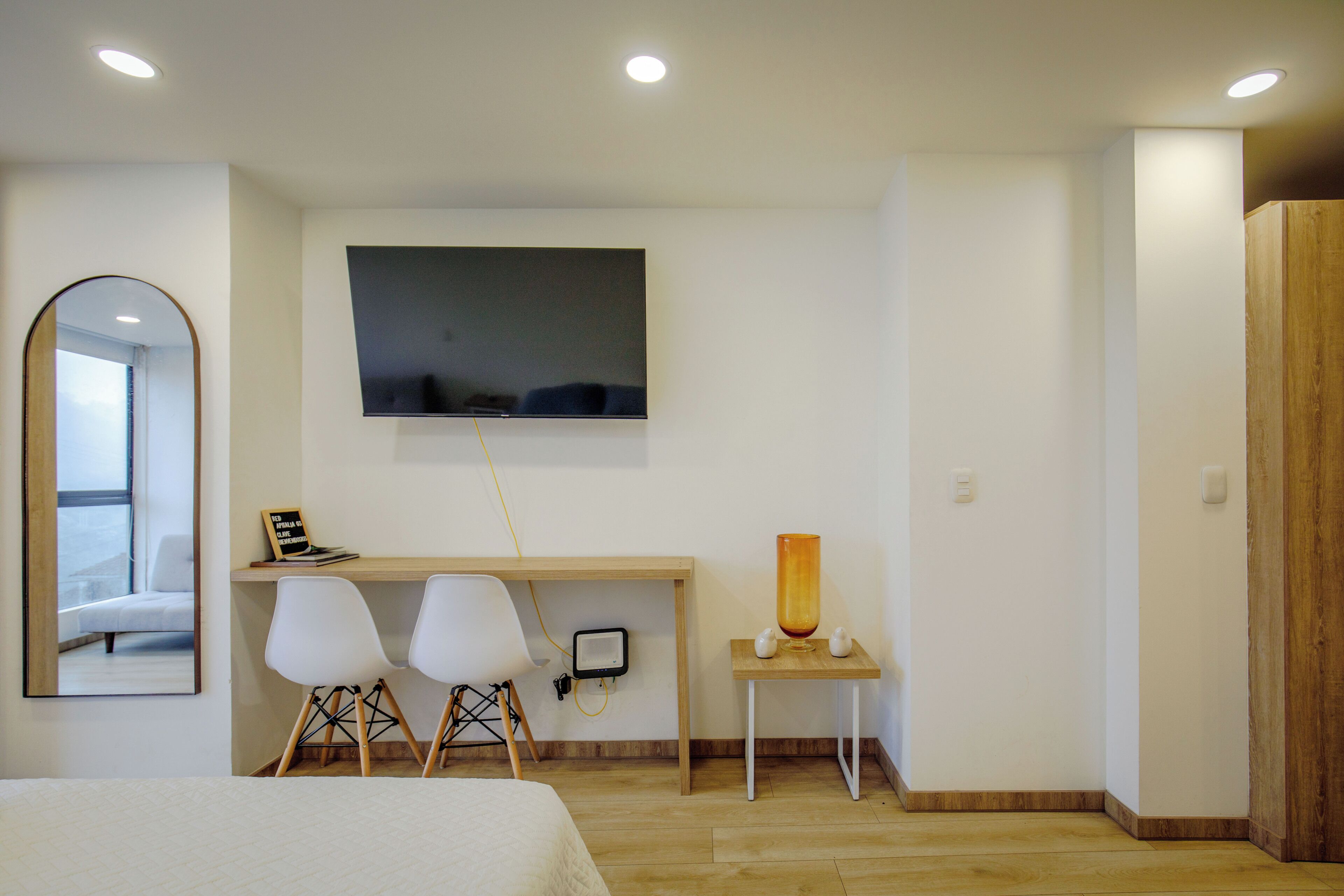 A modern bedroom with a bed, a desk with two chairs, a TV mounted on the wall, and a small table with a lamp.