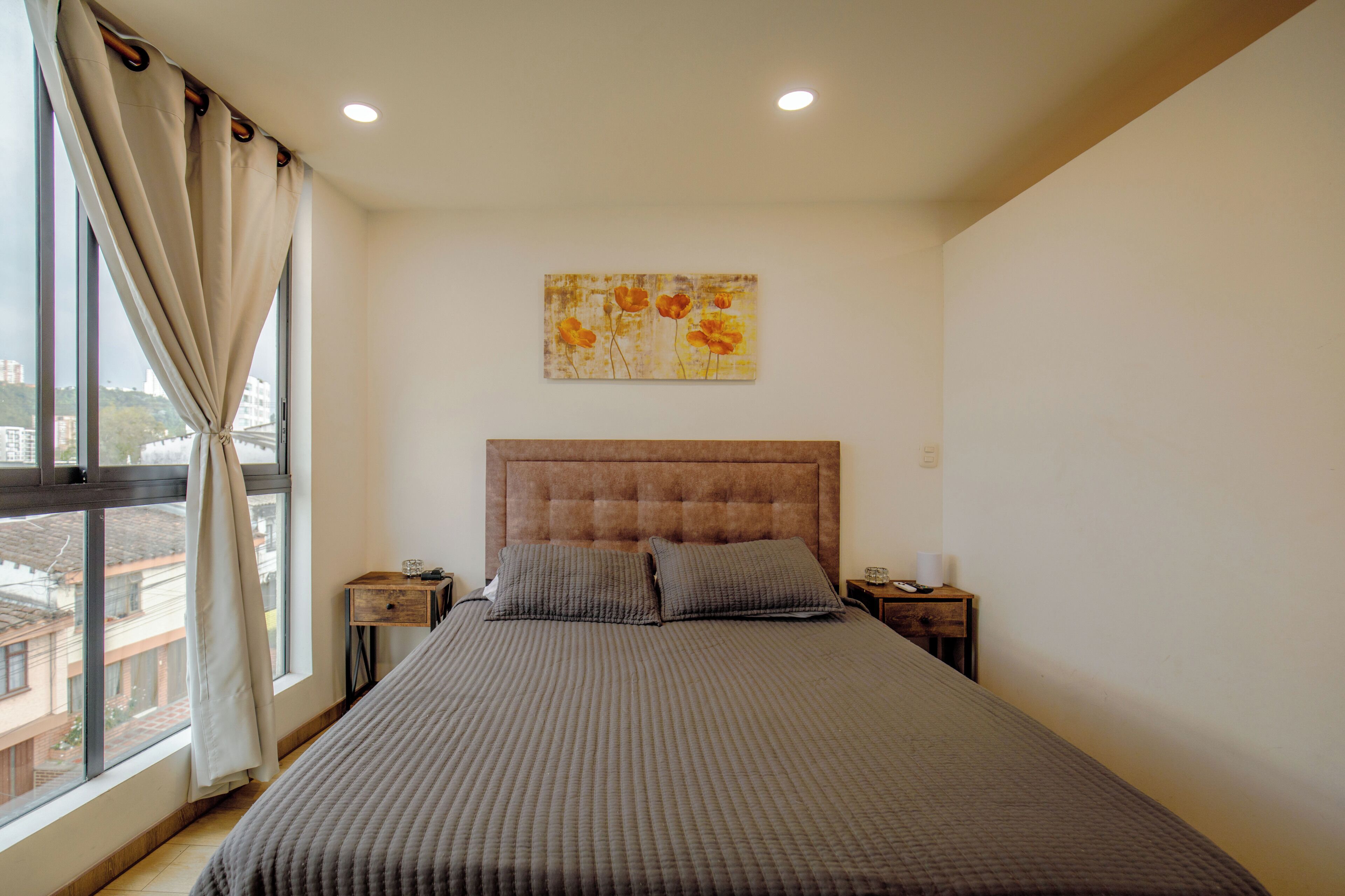 A bedroom with a large bed, a nightstand, a wall-mounted painting, and a window with curtains.