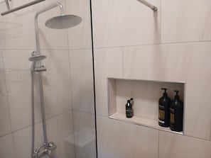 A shower with a handheld showerhead and a wall-mounted rain showerhead. There is a shelf with two bottles of shampoo and a bottle of lotion.