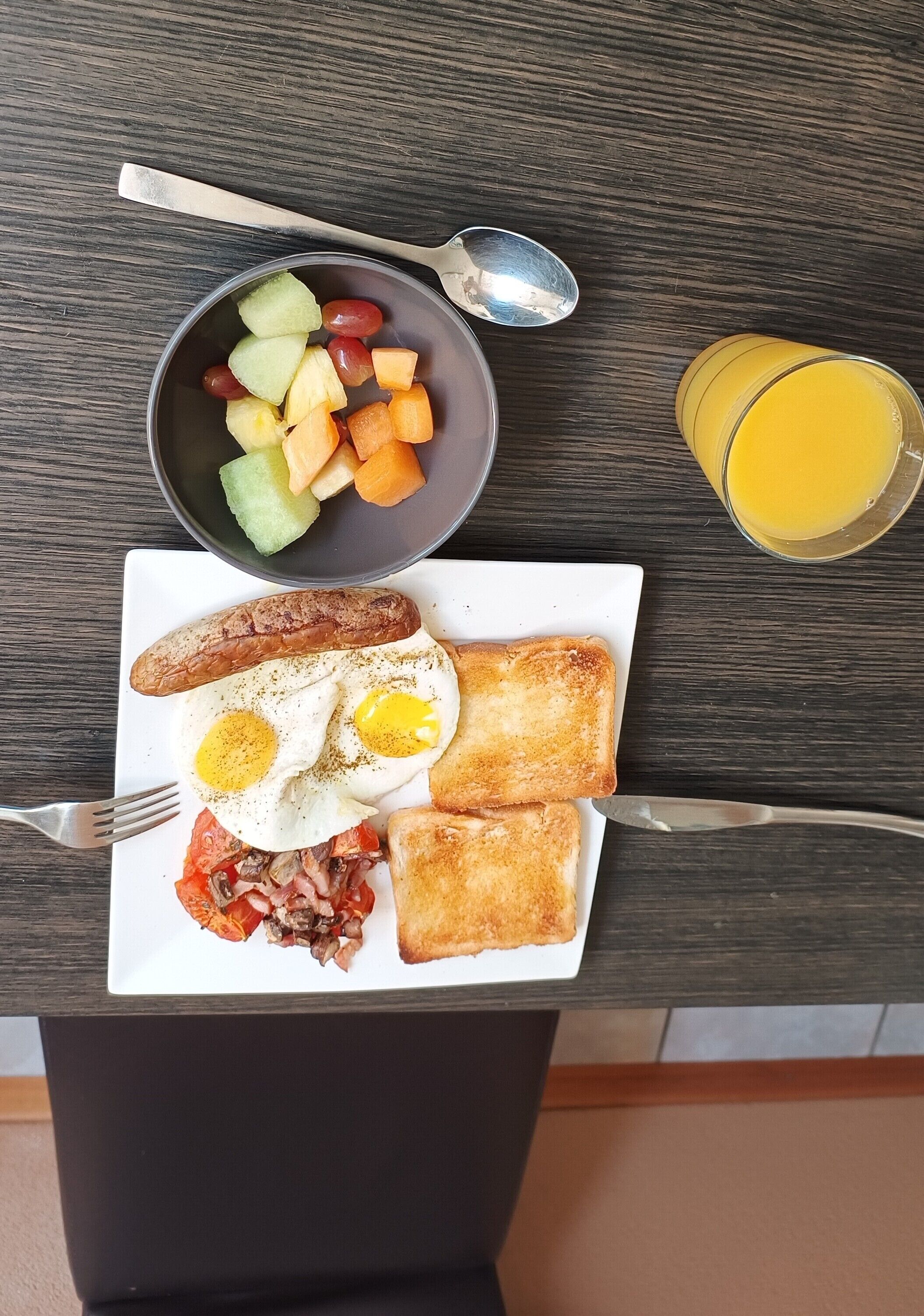 Une assiette de petit-déjeuner avec des œufs, des saucisses, des toasts et un bol de fruits, le tout posé sur une table.