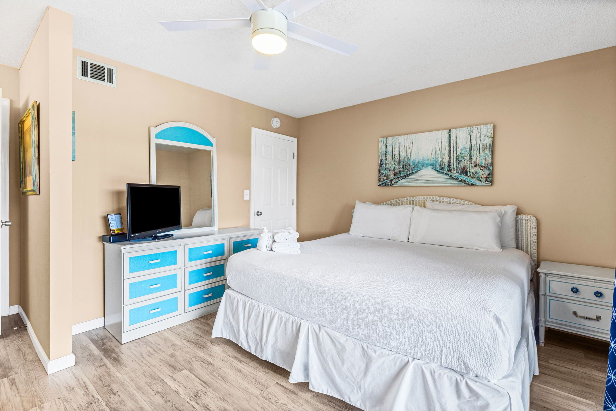 Une chambre à coucher avec un lit, une télévision, une commode, un tableau et un ventilateur de plafond.