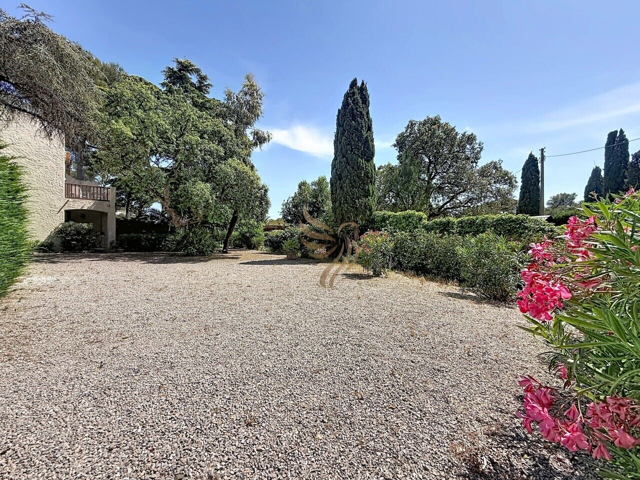 A gravel driveway leading to a building with a balcony, surrounded by trees and shrubs.