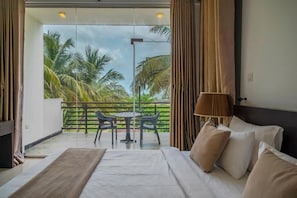 Ein Hotelzimmer mit Bett, Tisch, StĂŒhlen, einer Lampe und Blick auf Palmen durch eine GlastĂŒr.