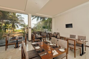 A dining area with multiple tables and chairs, large windows with a view of palm trees and a building, and a flat-screen TV mounted on the wall. - Beverly Beach Resort - Negombo (Negombo)
