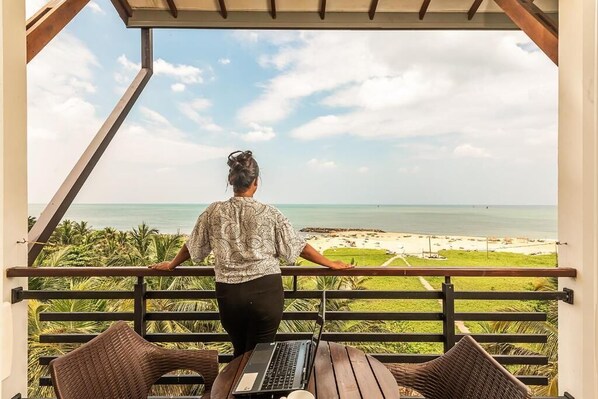 Eine Person steht auf einem Balkon mit Blick auf einen Strand, auf einem Tisch steht ein Laptop.