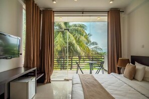 A bedroom with a bed, a TV, a balcony with a table and chairs, and a view of palm trees. - Beverly Beach Resort - Negombo (Negombo)