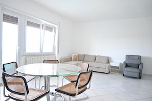 Un soggiorno moderno con un tavolo da pranzo con piano in vetro, sedie in vimini, una poltrona grigia, un divano e un tavolino bianco.