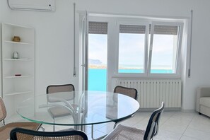 Habitació amb taula i cadires de vidre, radiador blanc, sofà blanc i vista al mar per la finestra.
