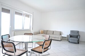 Una sala moderna con una mesa de comedor de vidrio, sillas de mimbre, un sillón gris, un sofá y una mesita auxiliar blanca.