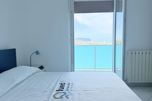 Un dormitorio con cama, mesita de noche, lámpara, ventana y vista al mar y una tierra distante.