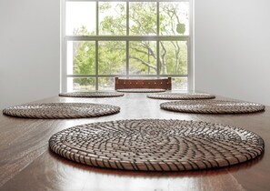 Mesa de comedor con individuales tejidos y un banco frente a una ventana con vistas a árboles.