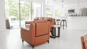Ein modernes Wohnzimmer mit einer Couch, einem Esstisch mit Hocker, einer Küchenzeile mit Geräten und großen Fenstern.