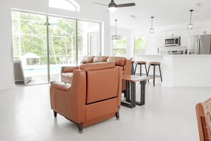 A modern living room with a sofa, a dining table with stools, a kitchen area with appliances, and large windows.