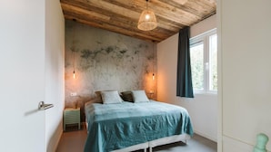 Un dormitorio con cama, techo de madera, dos luces fijadas a la pared, una ventana con cortinas y una mesita de noche pequeña.