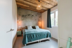Ein Schlafzimmer mit Bett, Holzboden, zwei Wandleuchten, einem Fenster mit Vorhängen und einem kleinen Nachttisch.