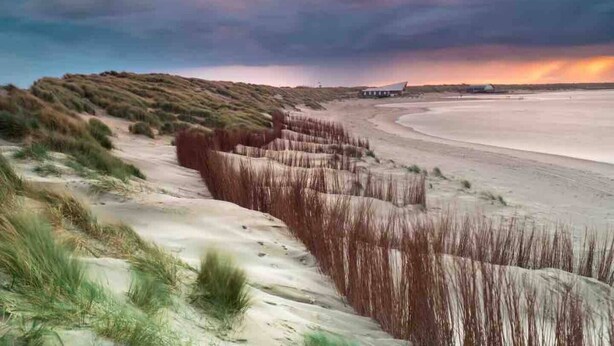 Paisaje costero con dunas, una playa y un atardecer.