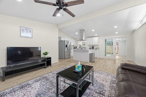 Un salon moderne avec un téléviseur à écran plat, une table basse en verre, un canapé en cuir marron, un ventilateur de plafond et une cuisine en arrière-plan.