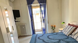 Een slaapkamer met een bed, een radiator, een televisie, een raam met gordijnen en een bloevas.