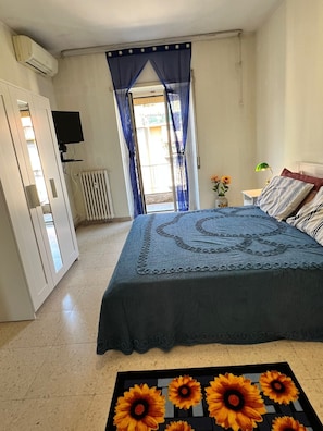 Ein Schlafzimmer mit Bett, Heizkörper, Fernseher, einem Fenster mit Vorhängen und einer Blumenvase.
