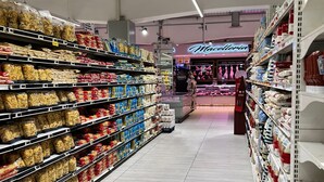 Een gangpad met pastasaus en blikken conserven op planken, een bakkerijgedeelte op de achtergrond, en een betegelde vloer.