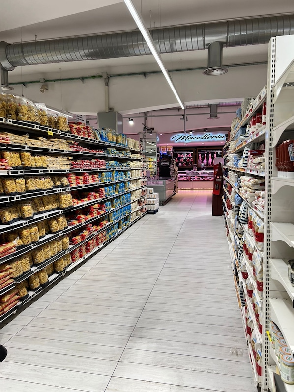 Ein Gang mit Nudeln und Konserven auf Regalen, im Hintergrund eine Bäckereiabteilung und ein gefliester Boden.
