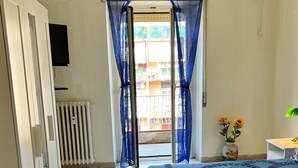 Een slaapkamer met een bed, een radiator, een televisie, een raam met blauwe gordijnen en uitzicht op de omgeving.
