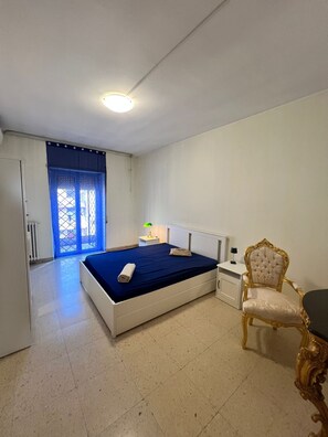 Un dormitorio con cama, una silla, una mesita de noche, una lámpara y una ventana con cortinas azules.