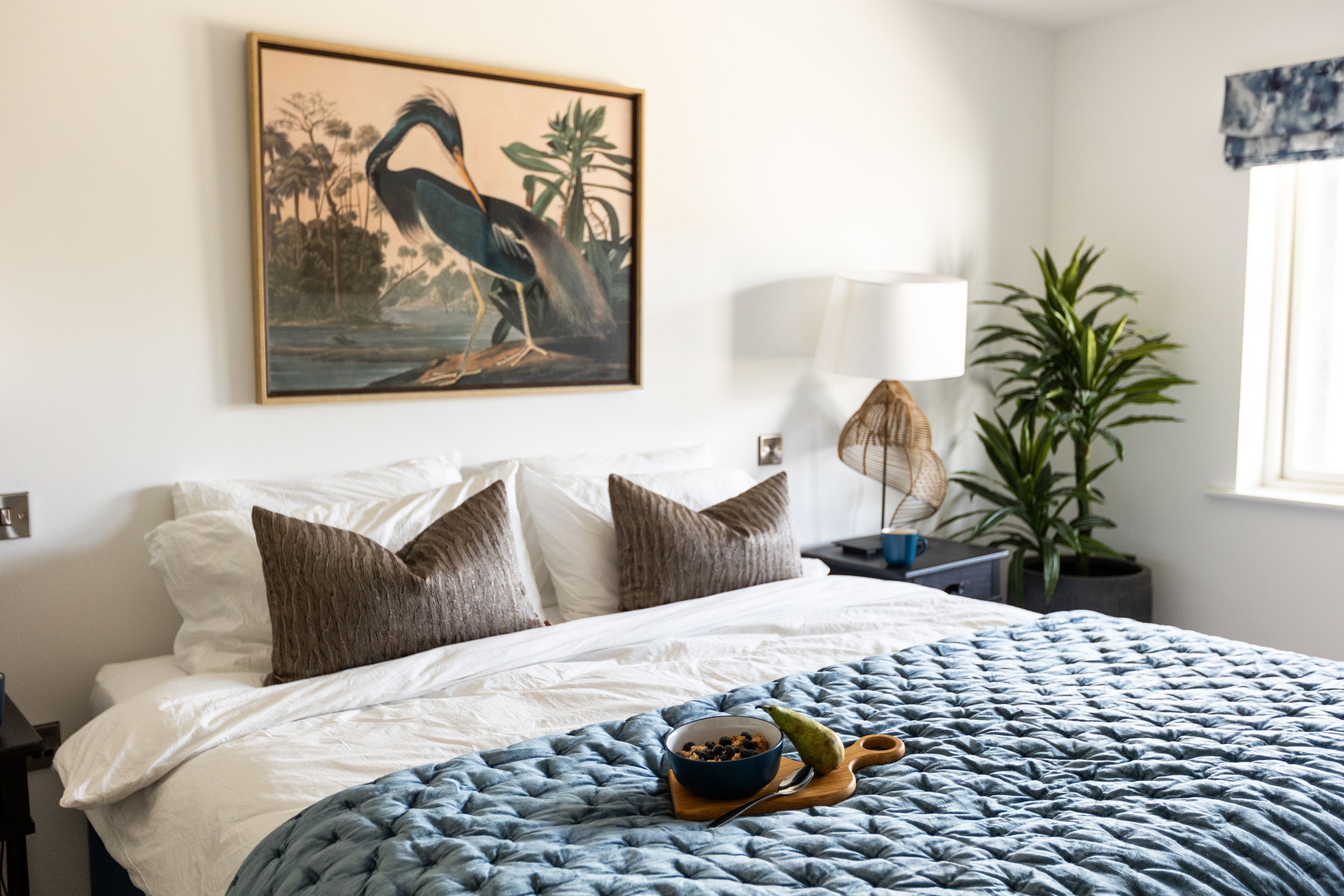 Una camera da letto con un letto, cuscini, un comodino con una lampada, una pianta e un quadro appeso al muro.