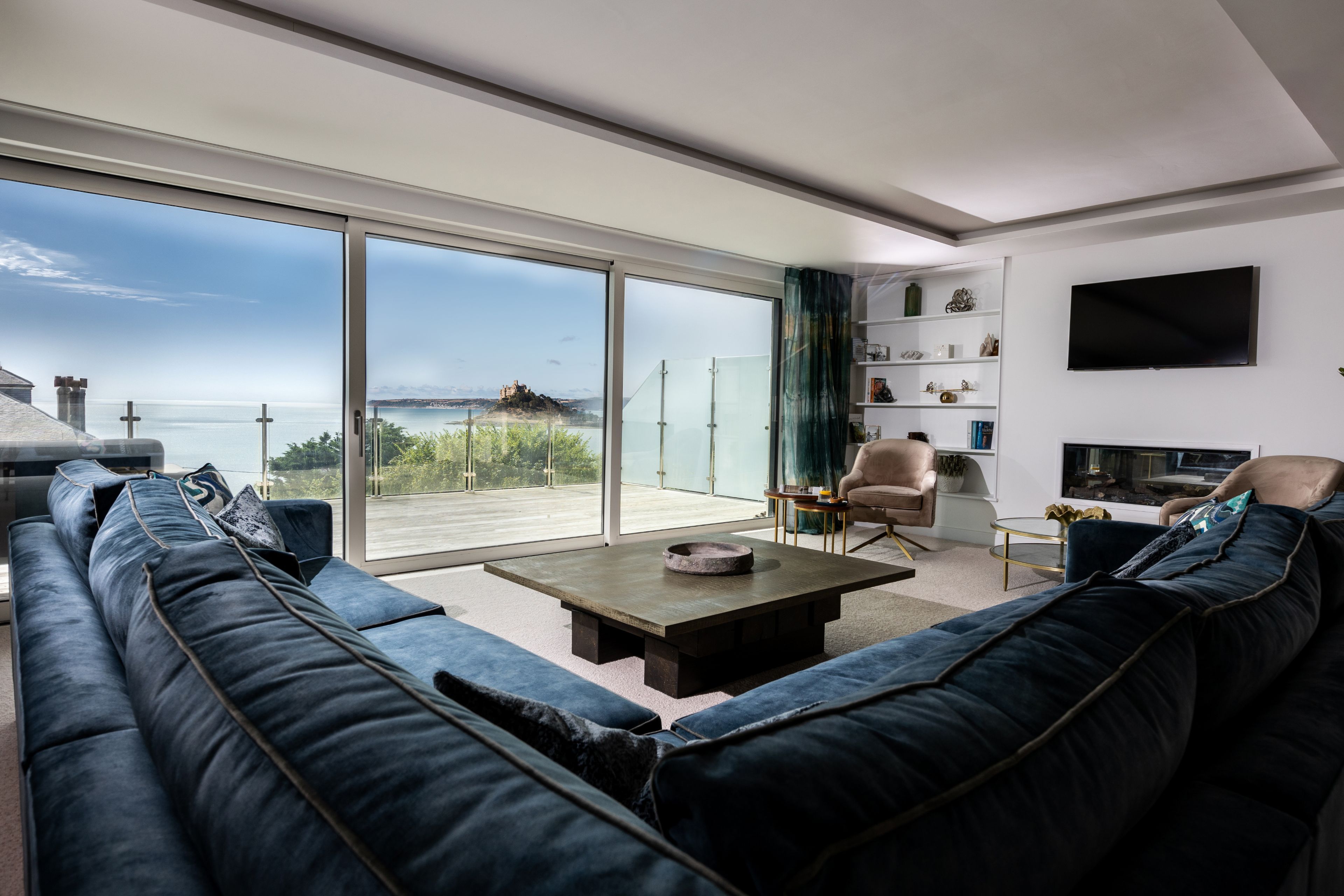 Een moderne woonkamer met een grote bank, een salontafel en een televisie. Er is een schuifpui naar een balkon met uitzicht op zee.