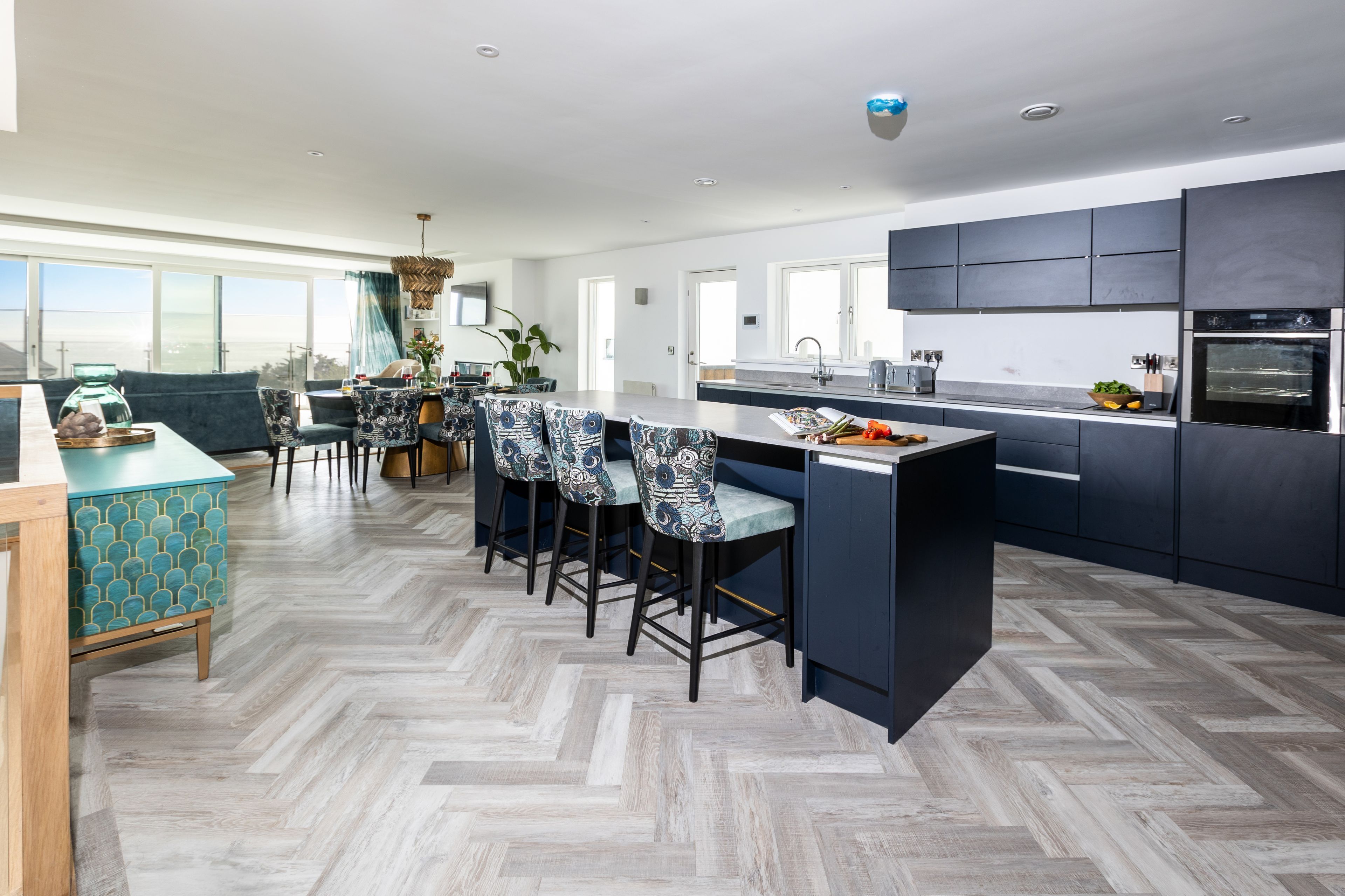 A modern kitchen with dark cabinetry, a herringbone floor pattern, and a dining area with patterned chairs.