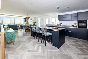 A modern kitchen with dark cabinetry, a herringbone floor pattern, and a dining area with patterned chairs. - Stunning Family Home Overlooking St Michaels Mount with hot tub (Marazion)