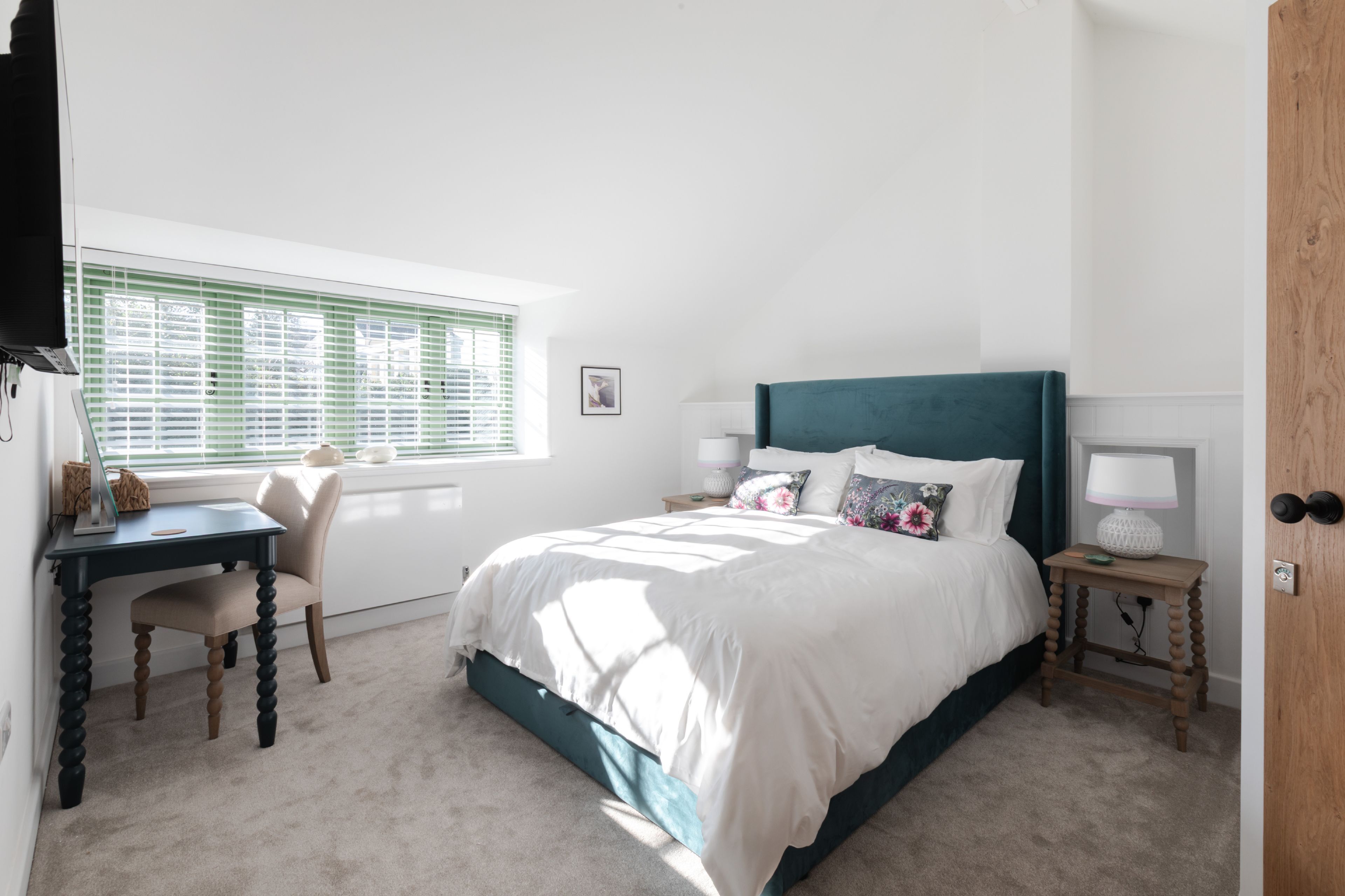 A bedroom with a bed, a desk, a chair, a window with blinds, and a door.