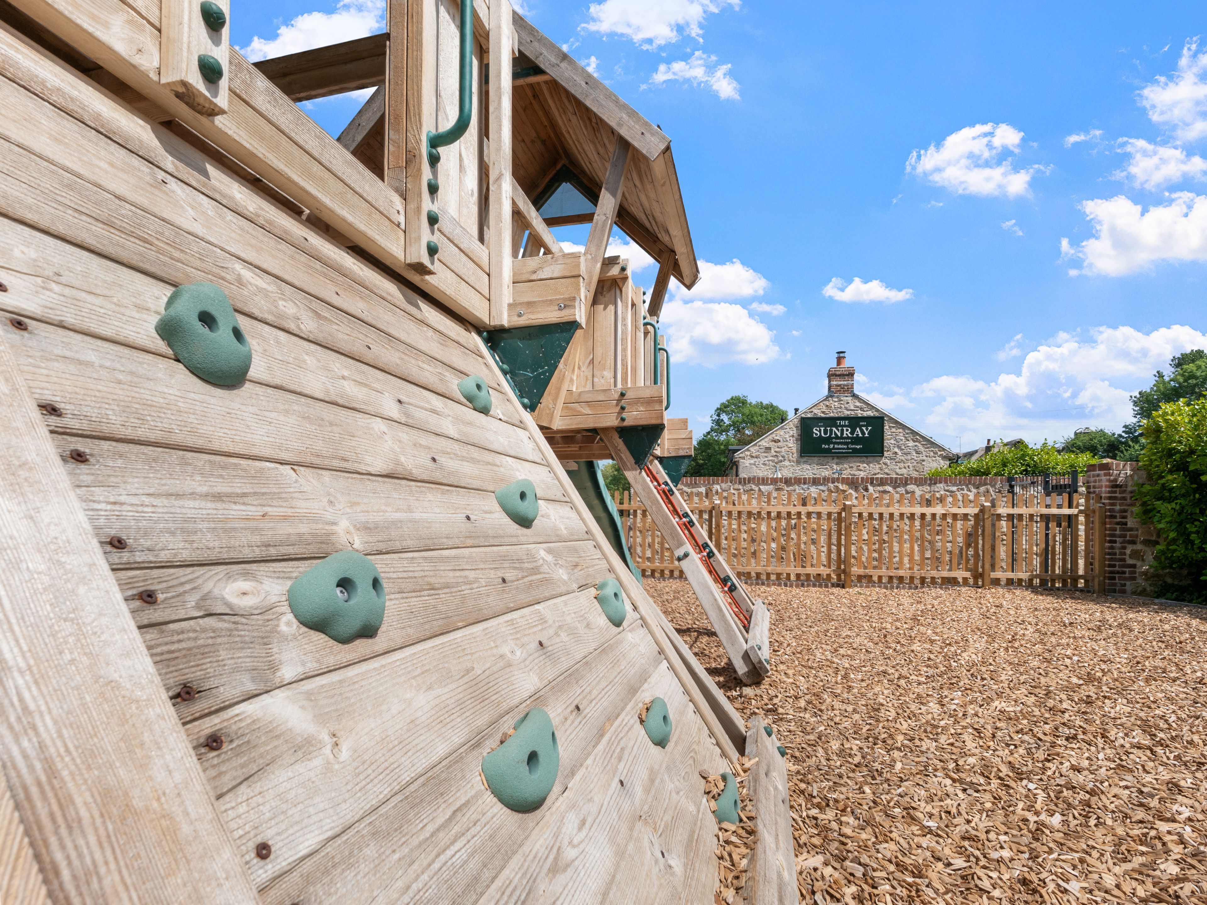 A wooden climbing wall with handholds, a wooden slide, and a wooden fence.
