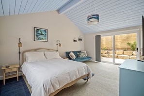 Ein Schlafzimmer mit Bett, Nachttisch, Lampe, Gemälde und Blick ins Freie durch eine Schiebetür.