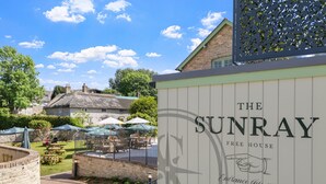 Une enseigne pour le « Sunray Free House », avec un logo, une indication de la direction de l’entrée et des places assises à l’extérieur, sous des parasols.