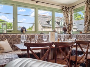 A dining area with a table set for six, a sofa, and a view of houses through large windows. - Wood-Fired Hot Tub & Pub Couples Retreat (Osmington, Weymouth)