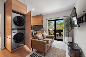 A modern apartment with a built-in washer and dryer, a sofa, a dining area with yellow chairs, and a balcony with a view.