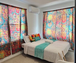A bedroom with a bed, colorful curtains, an air conditioner, and a pillow with text. - Casa Boricuá, Poblado Boquerón (Boquerón)