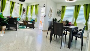 Amplia habitación con comedor, zona de estar con sofá y sillones, y un gran ventanal con cortinas verdes.