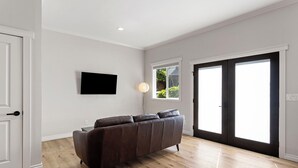 En moderne stue med en mørkebrun sofa, et fladskærms-tv ophængt på væggen og et stort vindue med udsigt til grønne omgivelser.