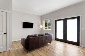 A modern living room with a dark brown sofa, a flat-screen TV mounted on the wall, and a large window with a view of greenery outside. - Peaceful 1 bed suite in fantastic location.  Close to everything. (Redmond)