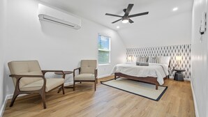 Een slaapkamer met een bed, twee stoelen, een plafondventilator, een airconditioner, een raam met zonwering en een kamer met een patroon op de muur.
