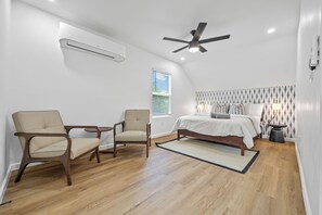 Ein Schlafzimmer mit einem Bett, zwei Stühlen, einem Deckenventilator, einer Klimaanlage, einem Fenster mit Jalousien und einer Wand mit Muster.
