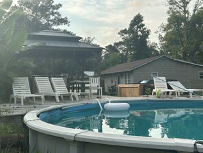 A swimming pool with lounge chairs and a covered area, surrounded by trees and a house.