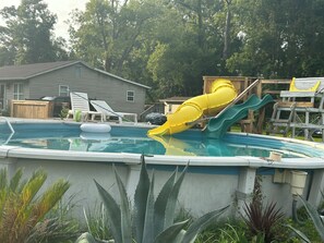 A swimming pool with a slide, surrounded by plants and a wooden deck.