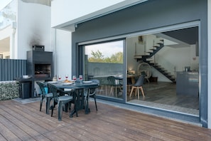Un espace repas extérieur moderne, avec une table et des chaises, un barbecue et une vue sur l’intérieur de la maison grâce à une porte vitrée.