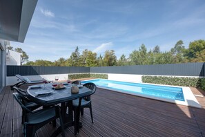 Un espace aménagé au bord de la piscine, avec une table et des chaises, une piscine, et une vue sur des arbres et une clôture.