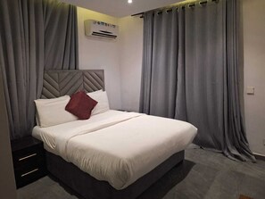 A neatly made bed with a headboard, grey curtains, and a small bedside table with drawers. - Sun Heaven Residence Lekki (Lekki)