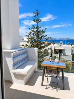 A balcony with a white sofa, a small table with a bottle, and a view of the sea and buildings. - Island Flavor - Naxos Seaview Retreat With Balcony (Naxos)