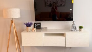 Ett modernt vardagsrum med en platt-TV som är monterad på en vit vägg, en golvlampa i trä, ett vitt konsolbord med dekorativa föremål och ett mörkt trägolv.